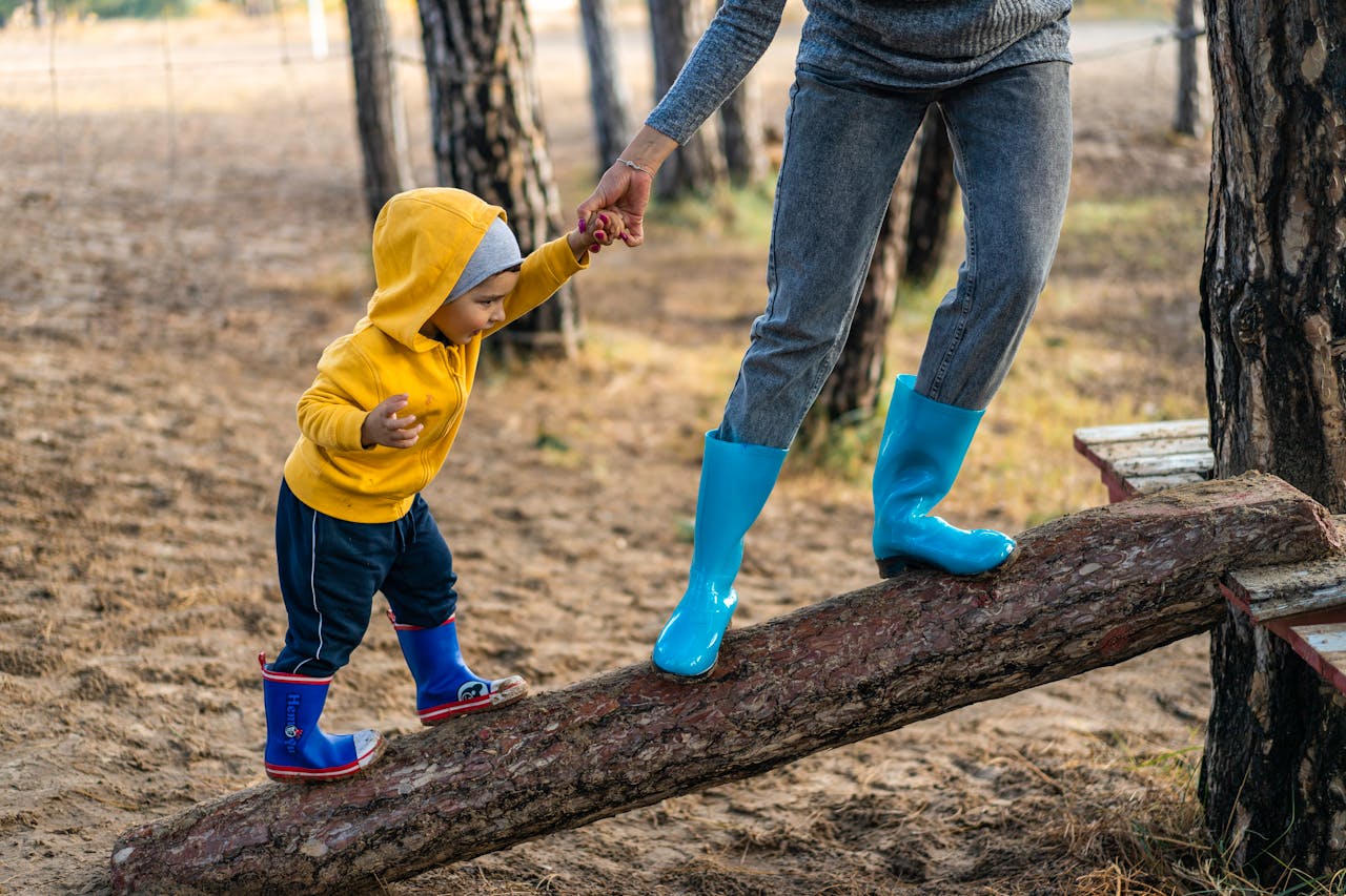 gallery-03 A woman helps her toddler walk on a log in a park, showcasing child support and family bonding.